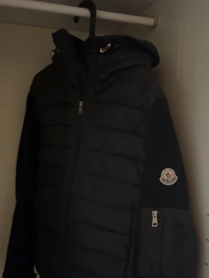 Moncler cardigan  - Säljer en svart dunjacka från Moncler med huva och dragkedja framtill. Jackan har Moncler-logga på ärmen och en praktisk ficka med dragkedja på ena ärmen. Klassisk quiltad design och sportig look, perfekt för kalla dagar.
