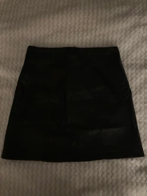 Svart skinnkjol med dragkedja - Snygg svart kort skinnkjol med rak passform och diskret dragkedja bak. Perfekt för dig som vill ha en stilren och edgy look. Kjolen är enkel att matcha och ger en cool vibe till din outfit.