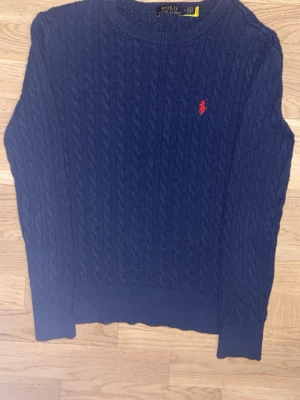 Mörkblå kabelstickad tröja Polo Ralph Lauren - Mörkblå kabelstickad tröja från Polo Ralph Lauren med rund halsringning och röd broderad logga på bröstet. Tröjan har långa ärmar och ribbade muddar vid ärmslut och nederkant. Perfekt för en klassisk och stilren look.