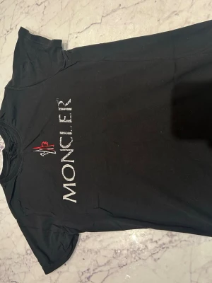 Svart Moncler t-shirt med logga - Snygg svart t-shirt från Moncler med stor vit logga och röd detalj på bröstet. Klassisk passform och rund halsringning. Perfekt till jeans eller shorts för en clean och stilren look.