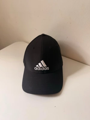 Svart keps från Adidas - Svart keps från Adidas med böjd skärm och justerbart spänne bak. Kepsen har klassisk sexpanelsdesign och vit broderad Adidas-logga framtill. Tillverkad i ett lätt och bekvämt material som passar perfekt till streetwear.