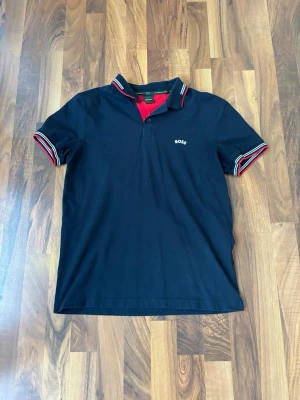 Marinblå pikétröja från BOSS - Snygg marinblå pikétröja från BOSS med röda och vita detaljer på krage och ärmslut. Tröjan har knappslå och logga på bröstet. Slim fit-modell i stretchigt material som sitter riktigt bra.