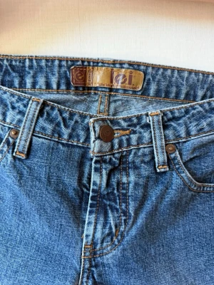 Blå jeans med snörning i benen - Unika blå jeans med snygg snörning längs benen. Klassisk femficksmodell med knappgylf och bälteshällor. Jeansen har en cool vintagekänsla och är tillverkade i slitstark denim. Perfekta för dig som vill sticka ut med en personlig stil.