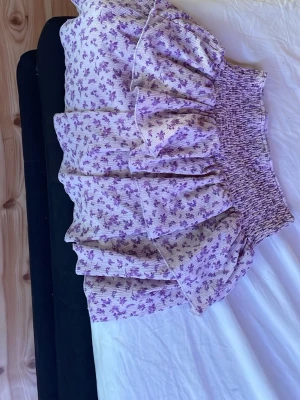 Ljuslila blommig volangkjol - Supersöt kort kjol i ljuslila med små blommor och volanger. Kjolen har bred resår i midjan för skön passform och är gjord i ett mjukt, lätt tyg som passar perfekt till varma dagar.