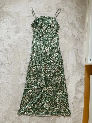Grön blommig långklänning L ZARA - Snygg långklänning i grönt med vita blommor och tunna axelband. Klänningen har en smickrande passform och ett somrigt blommönster som ger en fräsch look. Perfekt för varma dagar och speciella tillfällen.