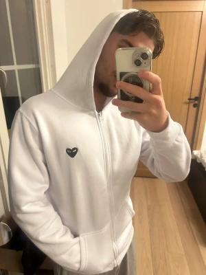 Comme des garçons - Säljer en vit hoodie med dragkedja och huva. På bröstet finns ett litet svart hjärta med ögon och på ryggen ett större svart hjärta med ögon. Hoodien har fickor framtill och ribbade muddar. Perfekt för en clean och stilren look. Den är i polyester material.
