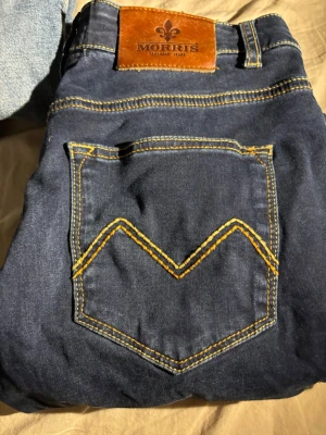 Morris Stevie Satin Jeans blå 29/30 - Mörkblå jeans från Morris i modellen Stevie Satin Jeans. Klassisk femficksdesign med kontrastsömmar i gult och läderpatch bak i midjan. Snygg V-formad söm på bakfickan. Perfekta för dig som gillar stilrena och tidlösa jeans.