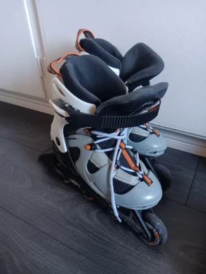 Gråa inlines med orange detaljer - Säljer ett par gråa inlines med svarta och orange detaljer. De har snörning, kardborreband och spänne upptill för extra stöd. Skalet är i plast och innerfodret är mjukt och vadderat för komfort. Perfekta för dig som vill cruisa runt i stan eller träna utomhus. Ställbara 35-38