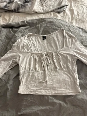 Vit croppad topp med knyt - Söt vit croppad topp med långa ärmar och en bred, fyrkantig urringning. Toppen har knyt framtill och lätt rynkade detaljer över bysten. Perfekt för dig som gillar en feminin och trendig stil.