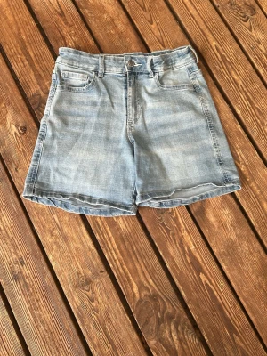 Ljusblå denimshorts med fickor - Säljer ett par ljusblå denimshorts med klassisk fyrfickig och knappgylf. Shortsen har en rak passform och kan vikas upp för en mer avslappnad look. Perfekta till sommaren och lätta att matcha med olika stilar. Har en liten fläck framvid men syns inte så tydligt