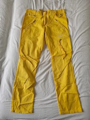 ÄKTA Y2K Gula T&T Cargobyxor, UNISEX / Streetwear - Perfekt för dig som vill sticka ut på stan i en helt unik streetwear-look. Chansen att du möter nåqon i samma par byxor är i princip noll.Storlek och Passform:Lappen säger XL, men då det är äldre vintagestorlekar stämmer det inte med dagens mått.  Se de exakta, manuellt mätta måtten nedan för perfekt passform: Midja (mätt platt): 42 cm (motsvarar ca storlek Medium för tjejer/ för killar W31–W32  Innerbensmått: 81 cm 