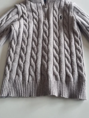 Grå stickad ralph lauren tröja - Snygg grå kabelstickad tröja med halv dragkedja och ribbade muddar. Tröjan har ett diskret broderat märke på bröstet och är gjord i ett mjukt, tjockt material. Perfekt för lager på lager och ger en klassisk look.