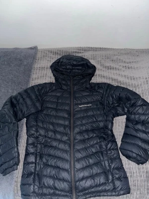 Svart dunjacka med huva Peak Performance - Snygg svart dunjacka från Peak Performance med huva och dragkedja framtill. Jackan har quiltad design och två sidofickor. Perfekt för kalla vinterdagar och riktigt lätt att bära. Klassisk och stilren modell som passar till det mesta.