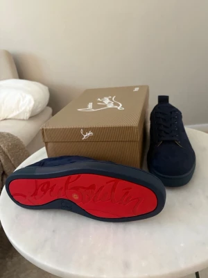 Marinblå sneakers från Louboutin - Snygga marinblå sneakers från Christian Louboutin i mocka med klassisk röd sula och diskreta svarta snören. Skorna har en låg profil och stilren design som passar perfekt till streetwear eller mer dressade outfits.