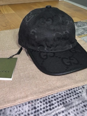 Svart gucci  keps  - Snygg svart keps från Gucci med det klassiska GG-mönstret över hela kepsen. Kepsen har böjd skärm med läderdetalj och är tillverkad i ett strukturerat tyg. Perfekt accessoar för en trendig och exklusiv look.