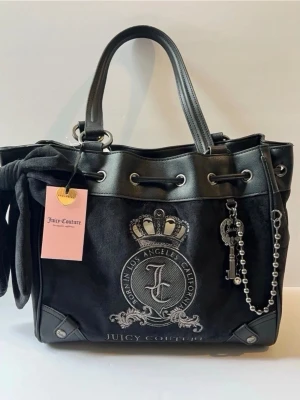 Svart handväska Juicy Couture daydreamer tote  - Snygg svart handväska från Juicy Couture med silverfärgade detaljer, emblem med krona och text, samt dekorativ nyckelberlock. Väskan har dubbla handtag, dragskostängning och en mjuk sammetsliknande yta med läderdetaljer. Perfekt för dig som gillar statement-accessoarer.