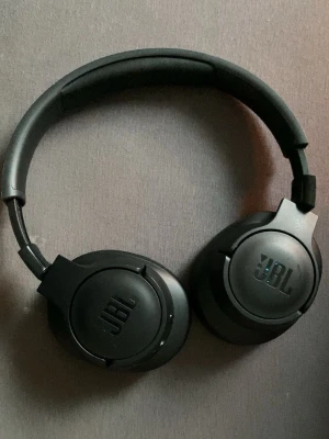 Svarta over-ear hörlurar från JBL - Snygga svarta over-ear hörlurar från JBL med vadderad bygel och mjuka öronkuddar. Hörlurarna har en stilren design i matt plast och är perfekta för musik, gaming eller videos. JBL-loggan syns tydligt på båda kåporna.