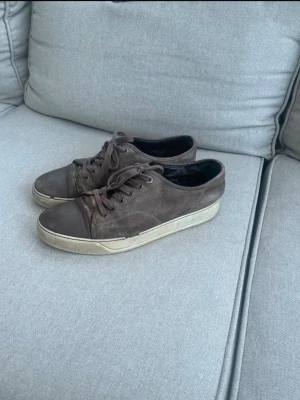 Bruna Lanvin sneakers i mocka - Snygga bruna sneakers från Lanvin i mjuk mocka med matchande bruna skosnören och tåparti i läder. Skorna har en vit, något smutsad sula och klassisk låg profil. Perfekta för dig som gillar stilrena och exklusiva sneakers.