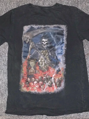 Svart Brave Soul t-shirt med dödskalletryck - Cool svart t-shirt från Brave Soul med stort tryck av dödskallen Grim Reaper på ryggen och mindre tryck med text på bröstet. T-shirten har rund hals och korta ärmar, perfekt för dig som gillar edgy motiv och streetwear.