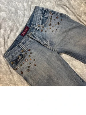 Low waist  jeans med nitar och bootcut - Säljer ett par ljusblå jeans från A ping med bootcut-modell och låg midja. Jeansen har coola nitar och metalldetaljer i olika former och storlekar framtill vid fickorna. Tillverkade i klassiskt denimtyg och perfekta för dig som vill sticka ut. De är ascoola men tyvärr lite för korta enligt mig, de går typ precis ner till marken för mig som är 166 cm. De har små rostfläckar längst ner som kommit från regnet, och de är uppsydda men inte klippta där nere. Midjemått: 38/39 cm Innerben: 72 cm
