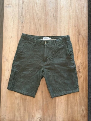Gröna linneshorts - Snygga gröna shorts i 100% linne med klassisk passform. Shortsen har två sidofickor fram, två bakfickor med knapp och bälteshällor i midjan. Perfekta för varma dagar och ger en avslappnad men stilren look. 