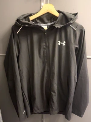 Svart vindjacka från Under Armour - Snygg svart vindjacka från Under Armour med huva och dragkedja framtill. Jackan har vita detaljer på axlarna och logga på bröstet samt stort Under Armour-tryck på ryggen. Tillverkad i lätt och vindtätt material, perfekt för blåsiga dagar.