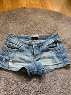 Blå jeansshorts från Wallflower - Säljer ett par blå jeansshorts från Wallflower med slitna detaljer och klassisk femficksdesign. Shortsen har låg midja och är gjorda i bomull med en snygg tvättad look. Perfekta för varma dagar och enkel att matcha med olika toppar. Finns ingen direkt storlek i dem men dom sitter som en S