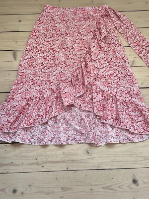 Blommig omlottkjol med volang - Söt blommig omlottkjol i rosa och vitt med volangdetaljer längs nederkanten och snörning i midjan. Kjolen har en luftig och flowy känsla, perfekt för varma dagar. Mönstret är småblommigt och kjolen är i en längre modell.