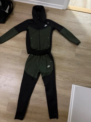 Nike grön och svart loungewear-set - Snyggt loungewear-set från Nike i mörkgrönt och svart. Setet består av en hoodie med dragkedja och fickor samt matchande byxor med resår i midjan. Båda delarna har Nike-logga framtill. Perfekt för chill eller träning.