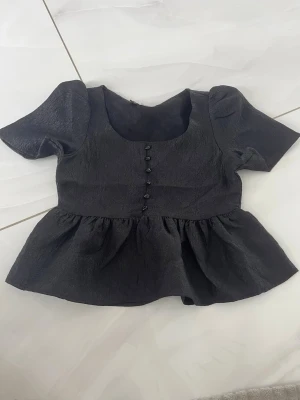 Svart blus med knappar och puffärm - Snygg svart blus med puffiga korta ärmar, markerad midja och volang nedtill. Framtill finns dekorativa knappar och tyget har en lätt glansig struktur. Perfekt för dig som vill ha en trendig och stilren topp med det lilla extra.