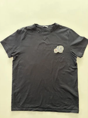 Svart t-shirt moncler - En svart t-shirt med rund hals och korta ärmar. På bröstet sitter en tydlig patch med ett märke och text. Storlek M! Bara att höra av dig vid minsta fundering😊
