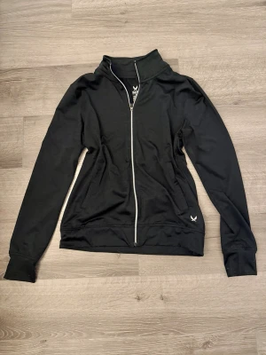 Svart träningsjacka från SOC XS - Snygg svart träningsjacka från SOC med hel dragkedja framtill och hög krage. Jackan har två sidofickor och en liten vit logga på nedre delen. Tillverkad i ett tekniskt material som andas och håller dig torr under träningen.