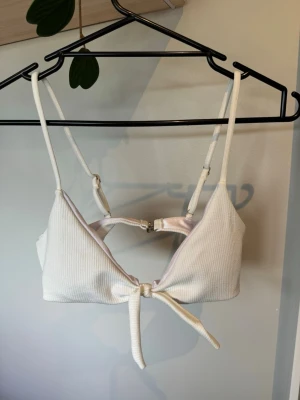 Vit ribbad bikiniöverdel med knyt - Snygg vit bikiniöverdel med ribbad struktur och justerbara axelband. Modellen har en dekorativ knytning framtill och metallspänne i ryggen. Perfekt för stranddagar och sommarhäng.
