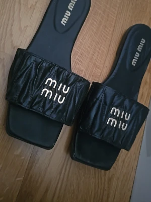 Svarta sandaler  - Säljer ett par svarta sandaler med quiltad rem och guldfärgad logga framtill. Sandalerna har fyrkantig tå och är tillverkade i vad som ser ut som läder. Perfekta för dig som vill ha en trendig och lyxig look.