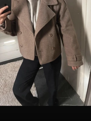 Kort brun ullrock med dubbelknäppning - Snygg kort brun rock i ullmix med bred krage och dubbelknäppning framtill. Rocken har raka ärmar med dekorativa spännen vid ärmsluten och en avslappnad passform. Perfekt för dig som vill ha en stilren och trendig look.