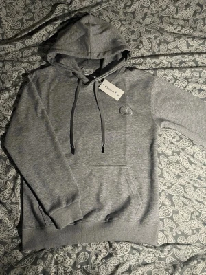 Grå hoodie - Snygg grå hoodie från med broderad CD-logga på bröstet. Hoodien har huva med dragsko, ribbade muddar och magficka. Tillverkad i mjukt bomullsmaterial som känns skönt mot huden. Perfekt för en avslappnad och trendig look.