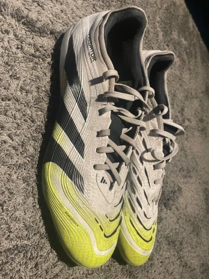 Adidas Predator fotbollsskor grå/gul - Snygga Adidas Predator fotbollsskor i grått med svarta detaljer och neon-gula partier framtill. Skorna har snörning, låg profil och är tillverkade i syntetmaterial för bra grepp och kontroll på planen. Perfekta för dig som vill sticka ut på matchen!
