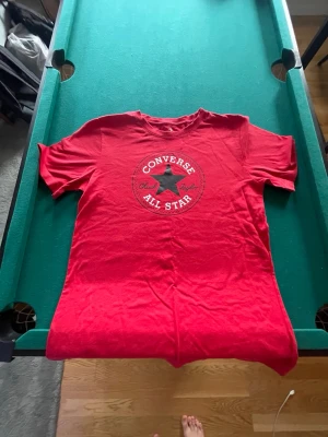 Röd Converse All Star t-shirt - Snygg röd t-shirt från Converse med det klassiska All Star-logotrycket i vitt och svart på bröstet. Modellen har rund hals och korta ärmar. Perfekt för dig som gillar streetwear och vill ha en ikonisk look.