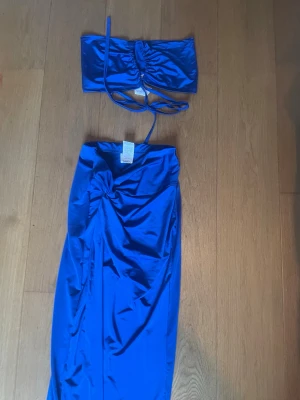 Blått set med kjol och topp - Snyggt tvådelat set i glansig blå satäng. Setet består av en bandeau-topp med smala knytband och en lång kjol med draperad detalj framtill. Materialet är slätt och skimrande, perfekt för dig som vill sticka ut.