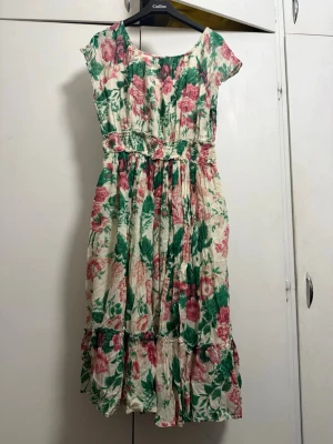 Blommig sommarklänning med volang - Söt blommig klänning i vitt med rosa och gröna detaljer. Klänningen har korta ärmar, markerad midja med smock och volangkant nertill. Perfekt för varma dagar och har en luftig, romantisk stil.