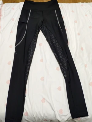 Svarta maya delorez  - Säljer ett par svarta sportleggings med hög midja och grå reflexdetaljer längs benen. Insidan av benen har ett mönstrat tyg med loggor och extra grepp. Perfekta för ridning Materialet är stretchigt och följsamt. några hål å så som jag har sytt ihop, haft de rätt länge nu men slutat med ridning 