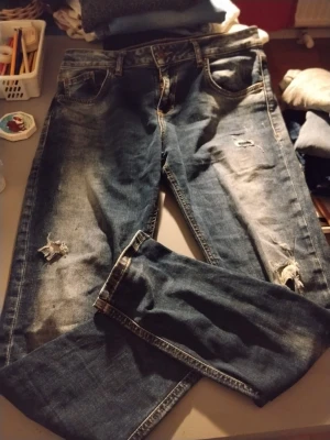 Blå slitna jeans från Amisu - Säljer ett par blå jeans från Amisu med snygga slitningar och rippade detaljer på benen. Klassisk femficksmodell med normal midja och raka ben. Jeansen är gjorda i denim och har en cool, avslappnad vibe.