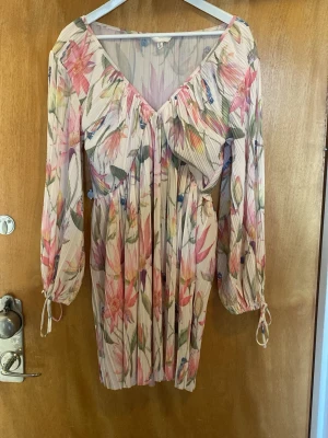 Blommig kortklänning från H&M - Säljer en blommig kortklänning från H&M med v-ringning och långa ärmar med knyt vid ärmslut. Klänningen har ett plisserat tyg i ljusa toner med inslag av rosa, grönt och beige. Perfekt för sommarens alla tillfällen!