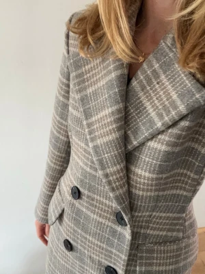 Grå-rutig dubbelknäppt kappa - Stilren grå kappa med rutigt mönster och breda slag. Dubbelknäppt framtill med svarta knappar och två fickor. Klassisk rak passform och tidlös design, perfekt för kyligare dagar. Materialet ser ut att vara ullblandning.