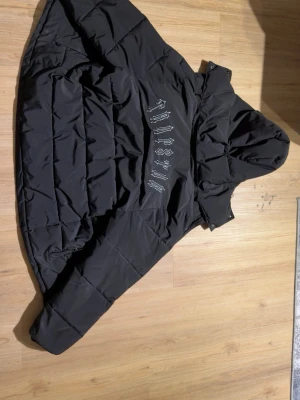 Svart pufferjacka från trapstar - Svart pufferjacka från Sixth June med stor huva och dragkedja framtill. Jackan har ett coolt tryck på ryggen i vit text och quiltad design. Perfekt för kalla dagar och riktigt street-inspirerad look.