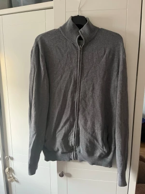 Grå ziptröja med hög krage - Säljer en grå ziptröja med hög ribbad krage och dragkedja hela vägen fram. Tröjan har långa ärmar, ribbade muddar och två fickor på sidorna. Perfekt för en avslappnad och clean look. Materialet känns mjukt och stretchigt.