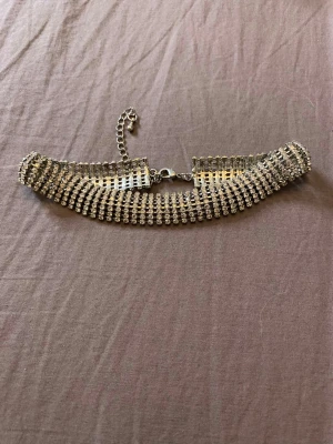 Strass choker - Snygg bred choker med flera rader glittrande strass i silverfärg. Halsbandet har justerbar kedja och karbinhake för enkel stängning. Perfekt för dig som vill sticka ut med ett statement-smycke. Materialet är metall och strass.