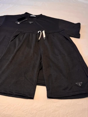 Svart loungewear-set från Prada - Snyggt svart loungewear-set från Prada med t-shirt och shorts. Setet har diskreta Prada-loggor på både tröjan och shortsen samt vita dragsnören i midjan. Tillverkat i ett mjukt och bekvämt material som passar perfekt för chill dagar hemma.