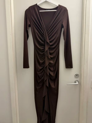 Brun långklänning med volang - Snygg brun långklänning med v-ringning och långa ärmar. Klänningen har en draperad volangdetalj framtill som ger en cool och trendig look. Perfekt för dig som vill sticka ut med en elegant och modern stil.