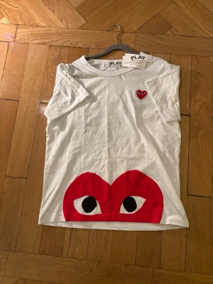 Cdg tisha  - Vit t-shirt från Comme des Garçons PLAY med det ikoniska röda hjärtmärket broderat på bröstet och ett stort rött hjärta med ögon tryckt längst ner. Klassisk passform och rund hals. Perfekt för dig som gillar streetwear och vill sticka ut.